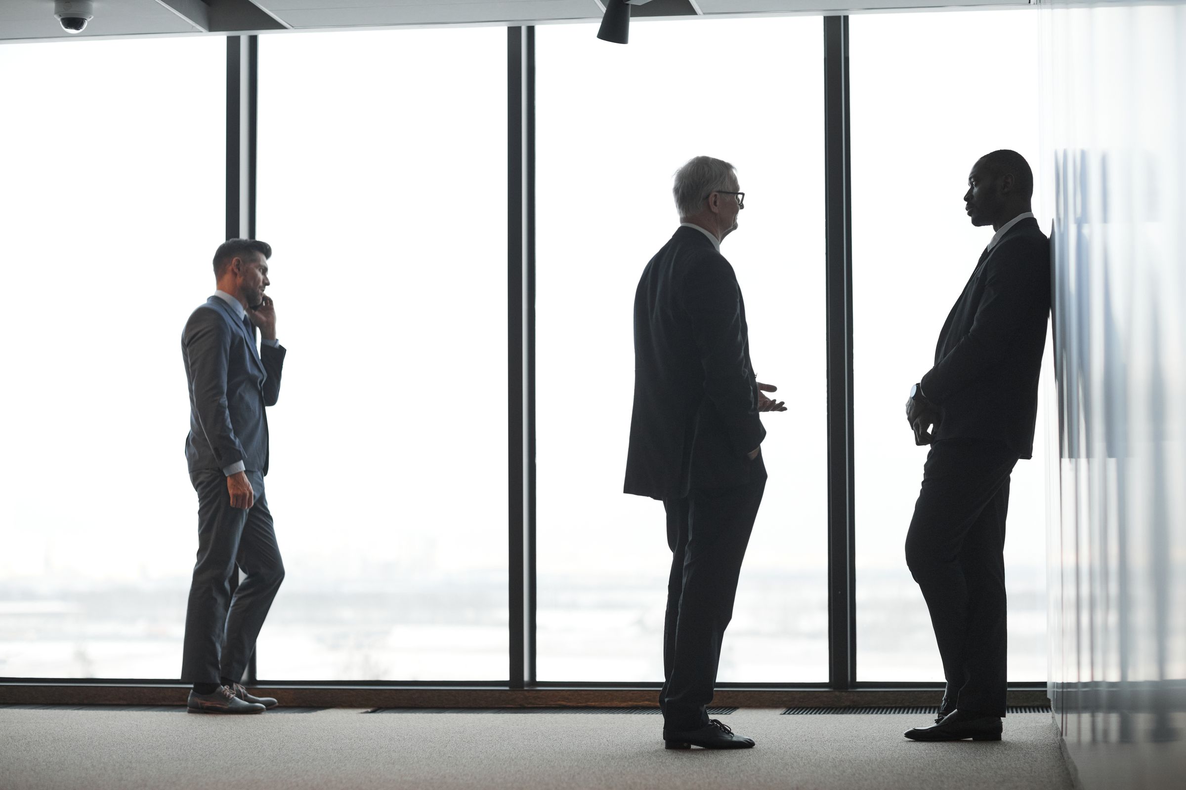 Business people backlit by tall office windows, one on a call while two others talk