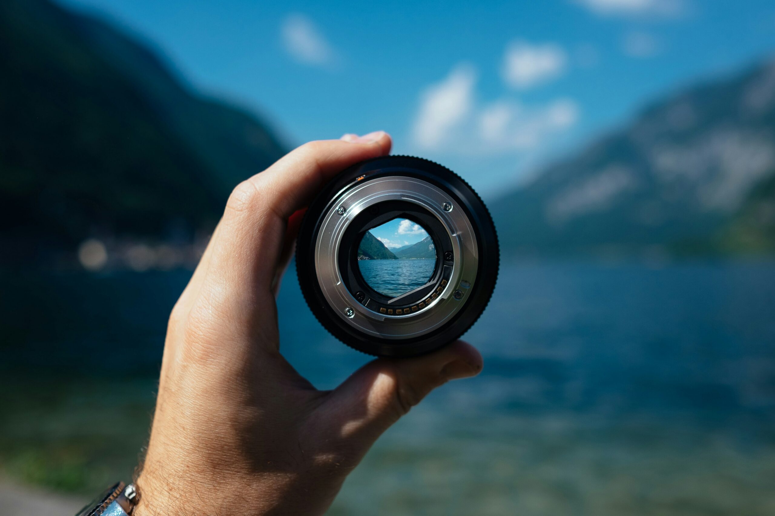 Hand holding a camera lens up to a lake and mountains